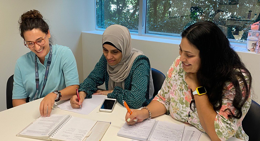 Three people are sitting at a table, writing in notebooks and using a smartphone, in a room with a window.