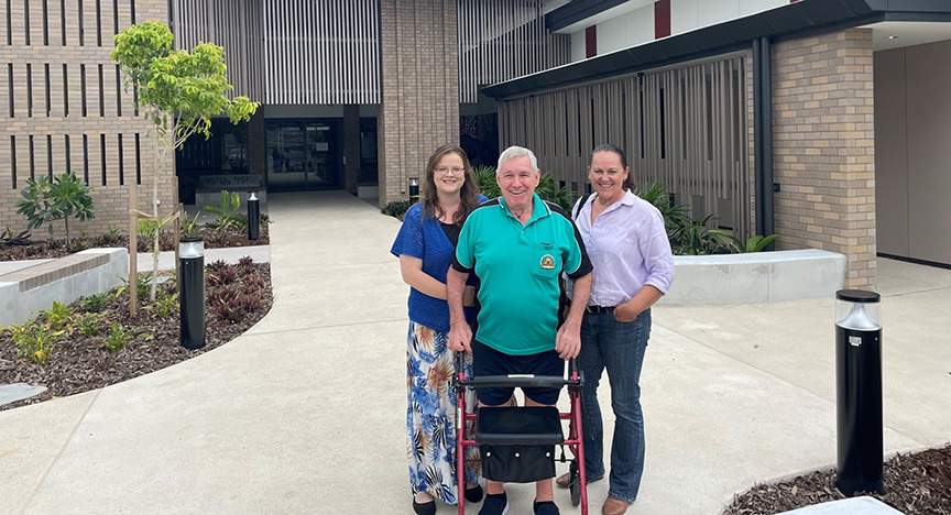 Three people stand outside a modern building, with one person using a walker. The setting appears to be a healthcare facility.