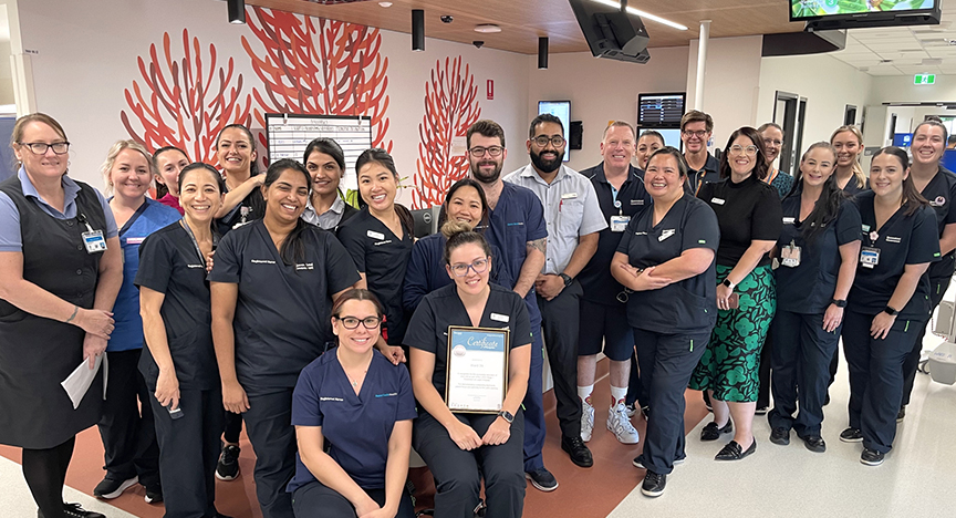 A group of healthcare professionals in uniforms pose together in a hospital corridor, with one person holding a certificate.