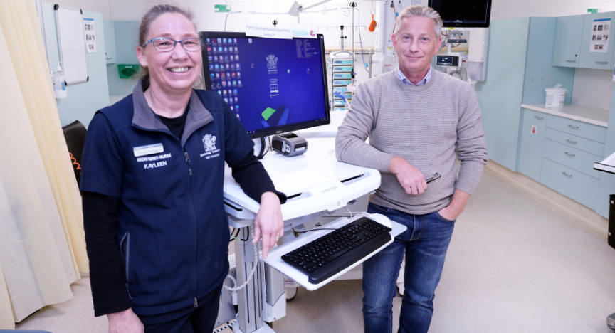 Two healthcare professionals stand beside medical equipment in a hospital room. One wears a uniform with a name badge.