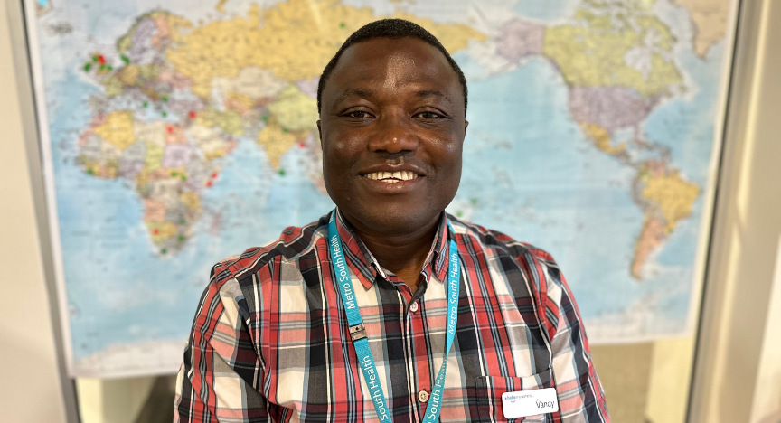 A person wearing a plaid shirt and a Metro South Health lanyard stands in front of a world map.