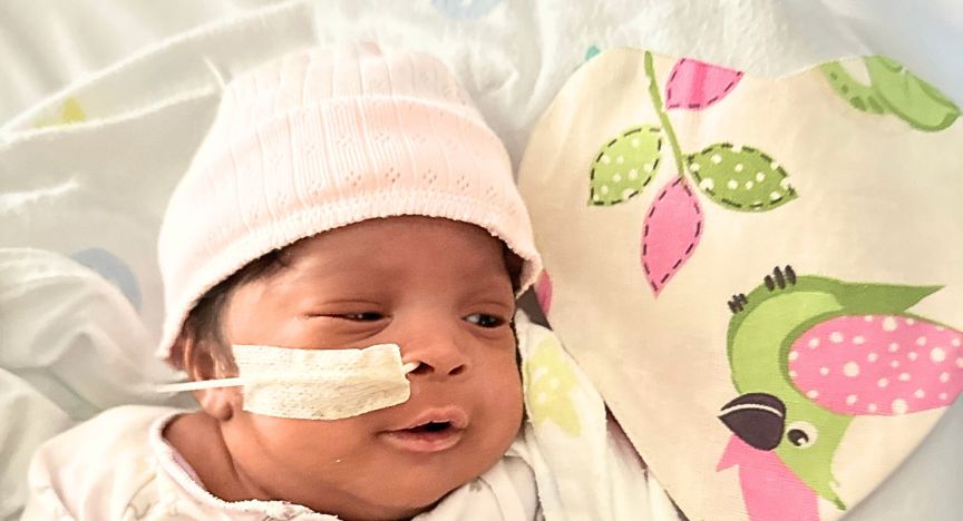 A baby wearing a pink hat lies next to a heart-shaped fabric with colorful bird and leaf patterns.