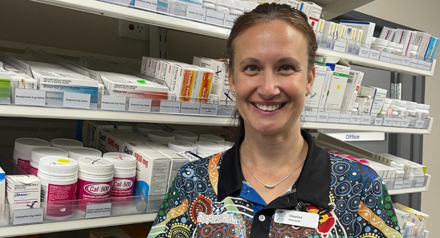 A close up headshot of a clinical pharmacist standing in front of a medication stand, wearing a First Nations polo shirt