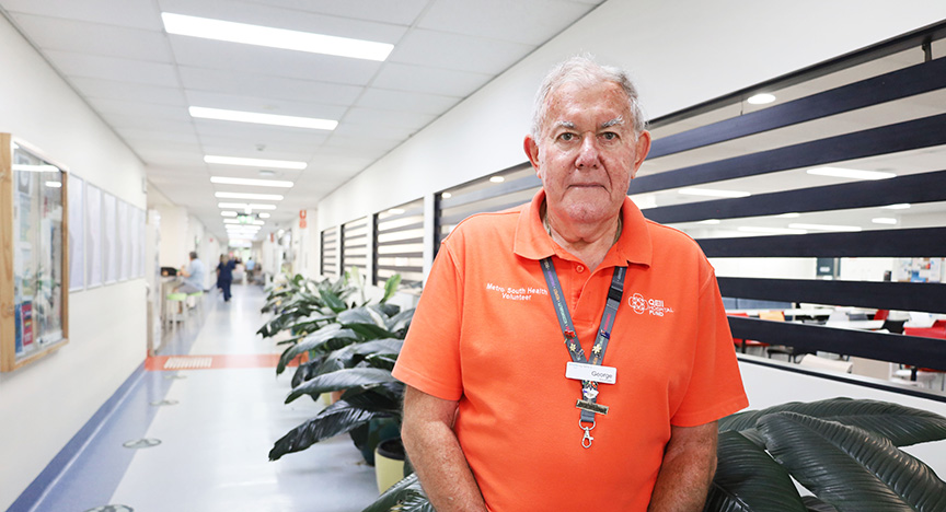 A hospital volunteer in an orange shirt stands in a hallway with plants and bulletin boards.