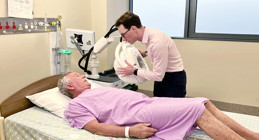 A patient in a hospital bed undergoes a brain scan using a specialized helmet operated by a healthcare professional.