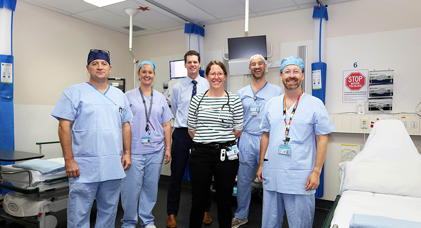 A group of healthcare professionals in a hospital room, standing together near medical equipment and a hospital bed.