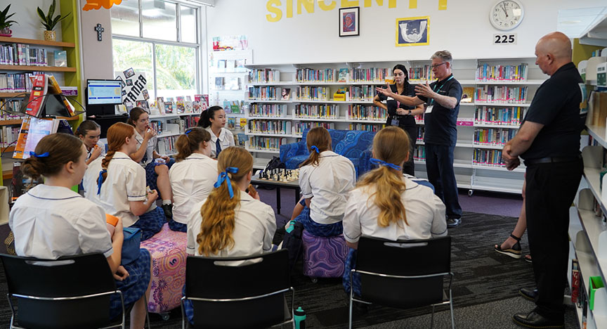 A group of students in a library listens to a presentation by two adults. The room has bookshelves, a computer, and a clock showing 2:25.