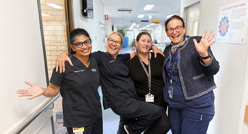 Four healthcare workers in uniforms pose cheerfully in a hospital corridor, celebrating an achievement.