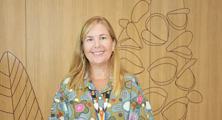 A person wearing a colorful shirt and a lanyard stands in front of a wooden wall with abstract line art.