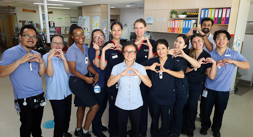 A group of healthcare workers in uniforms stand together in a hospital, forming heart shapes with their hands.