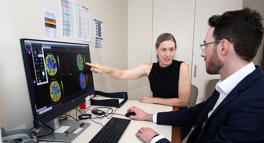 A person points at brain scan images on a computer screen while another person observes, likely discussing medical results.