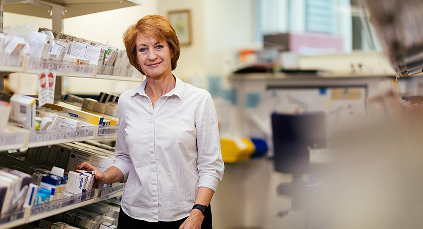 A pharmacist in a white shirt stands in front of shelves filled with various medications in a pharmacy.