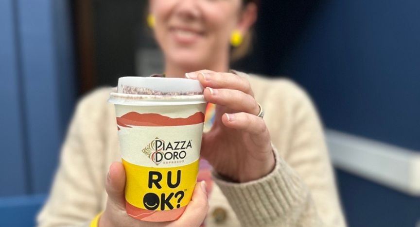 A person holds a Piazza D'Oro coffee cup with RU OK? written on it, promoting mental health awareness.