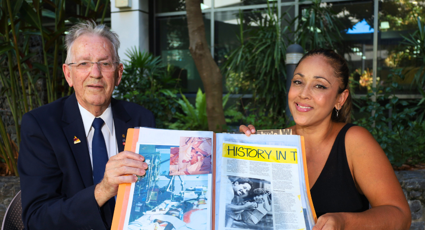 Professor Russell Strong and Rhonda Natera celebrate 40 years of liver transplants, holding a magazine filled with historic newspaper clippings at a table