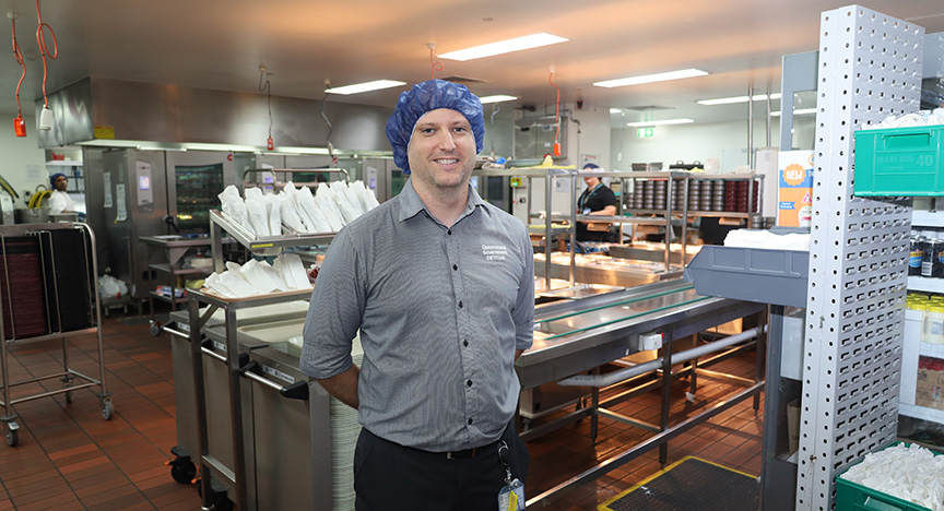 A person stands in a commercial kitchen with food preparation areas and equipment visible in the background.