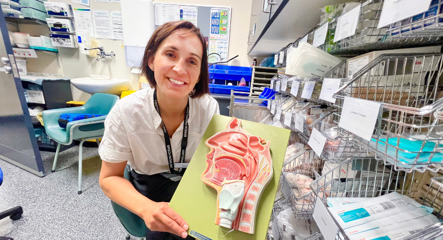 A person in a medical office holds a model of the human throat and vocal cords, surrounded by medical supplies and equipment.