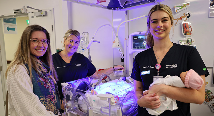 Two nurses and a visitor stand by a neonatal incubator with a baby receiving phototherapy. One nurse holds another baby.