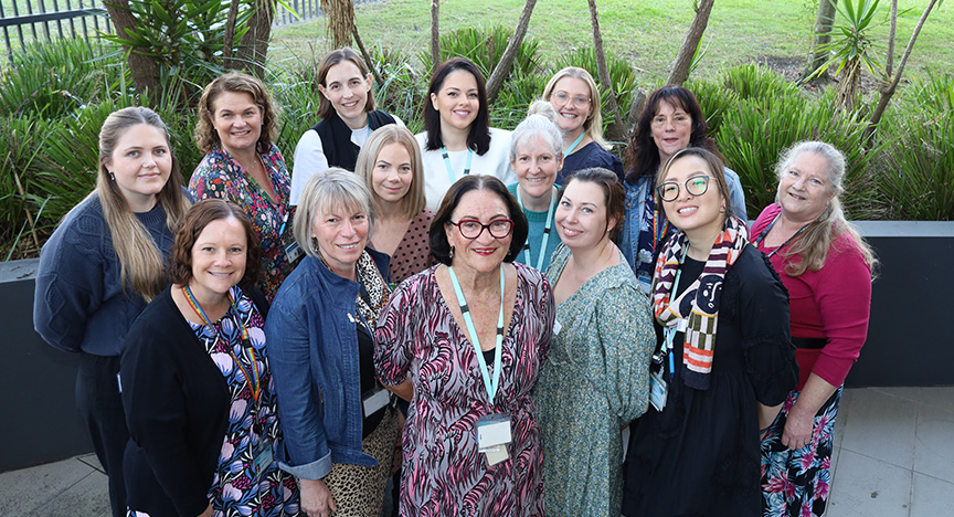 A group of people standing outdoors, some wearing lanyards, in front of greenery. They appear to be part of a professional or community event.