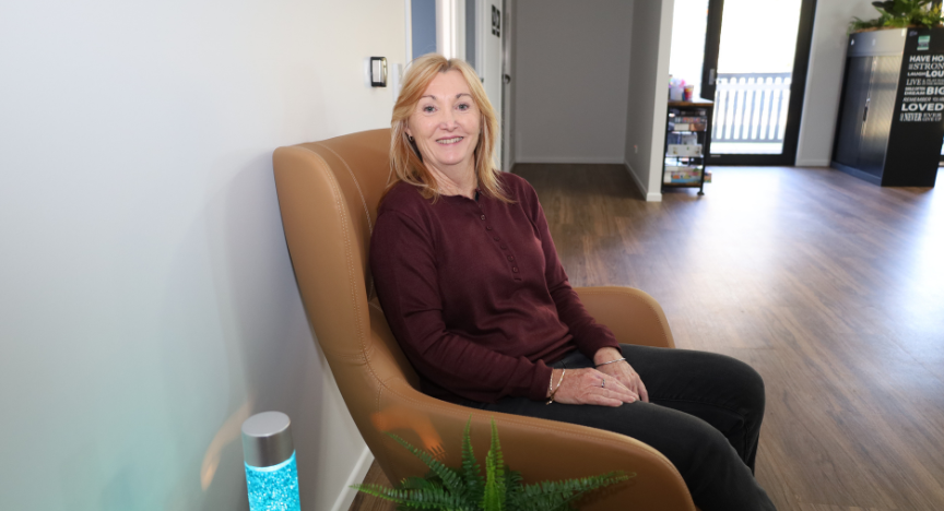 A person sits in a comfortable chair in a well-lit room with wooden flooring and a plant nearby.