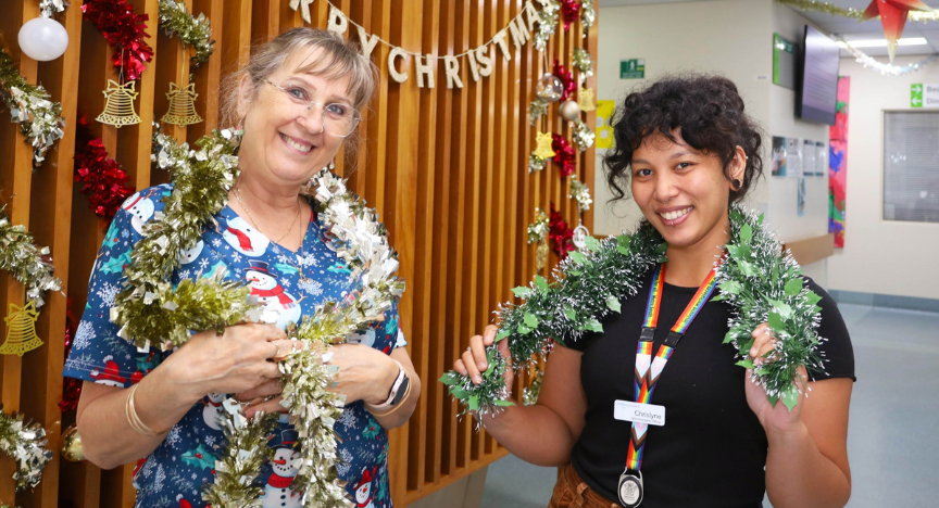 Two female mental health staff members standing in unit wearing Christmas tinsel around their shoulders with Merry Christmas sign in background