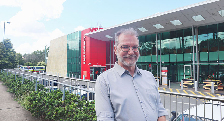 A person stands in front of the emergency entrance of a modern hospital building with glass windows and a red facade.