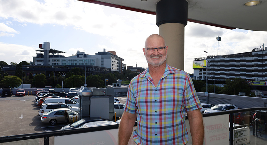 A person in a colorful plaid shirt stands on a balcony overlooking a parking lot and modern buildings in the background.