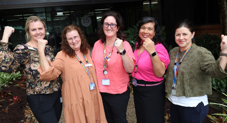 Five people standing outside a building, raising their fists in a gesture of strength and solidarity, wearing ID badges and colorful lanyards.