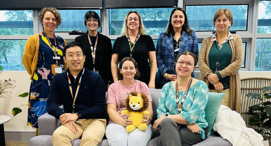 A group of eight people, some seated and some standing, pose indoors. One person holds a stuffed lion toy. They are in a room with large windows.