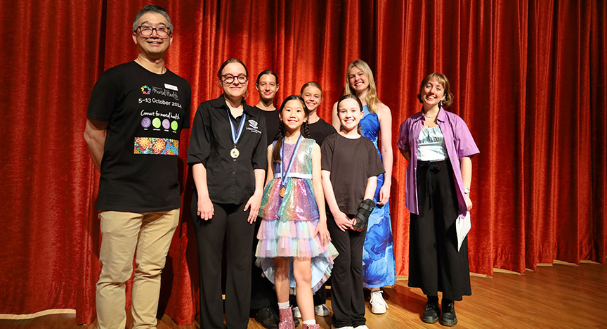 A group of people standing on a stage with a red curtain backdrop, some wearing medals and colorful outfits, likely at a mental health event.