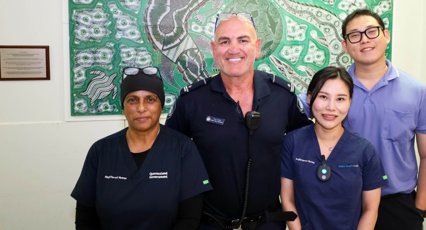 Four health staff standing in front of a large green and black Indigenous painting crafted by prisoners