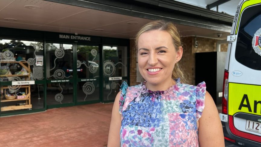 A healthcare worker in a floral top stands outside the entrance to Beaudesert Hospital. You can see part of an ambulance behind her.