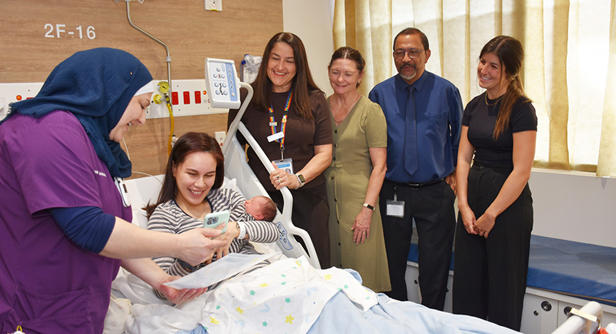 A healthcare worker assists a new mother with a mobile device while others stand by her hospital bed.