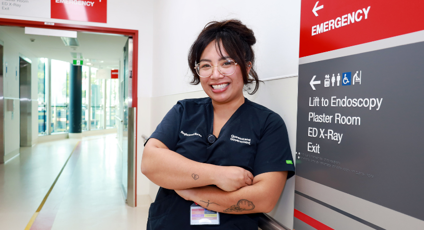 A healthcare worker stands in a hospital corridor near an emergency sign, wearing a uniform with Queensland Government and Registered Nurse badges.