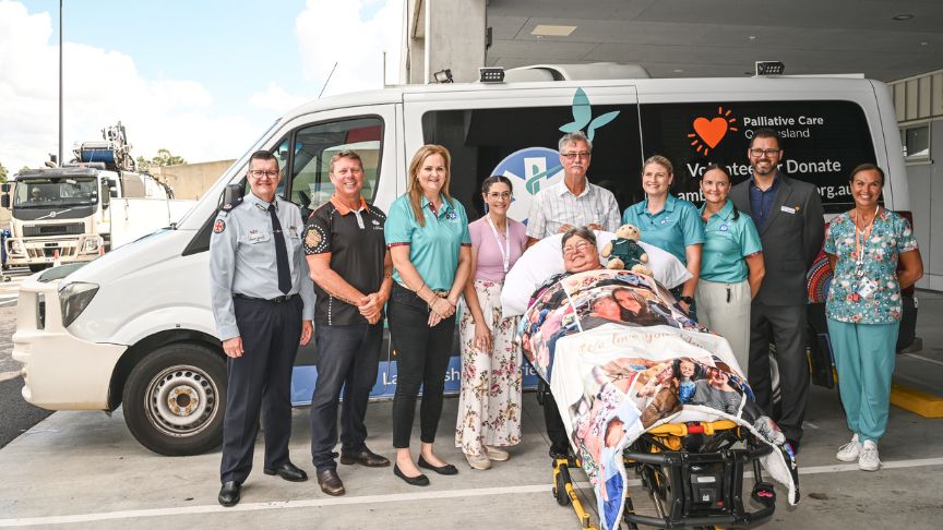 a large group of people stand in front of the Ambulance Wish QLD Amublance. In front of the group is a lady lying in a stretcher bed covered in a blanket that shows the faces of her grandchildren