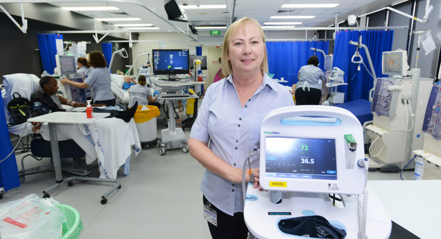 A healthcare worker stands in a dialysis unit with medical equipment in the foreground and patients receiving treatment in the background.