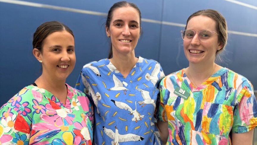 a group of nursing educators stand in front of a blue wall in brightly coloured srubs. One of them is wearing glasses and all 3 have their hair in a pony tail.