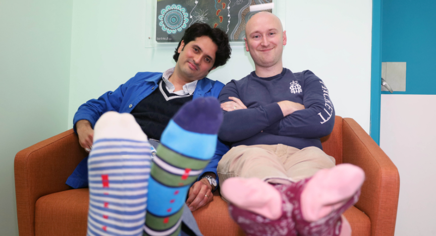 Two people sitting on an orange couch, showing off their colorful socks. A First Nations artwork is visible on the wall behind them.