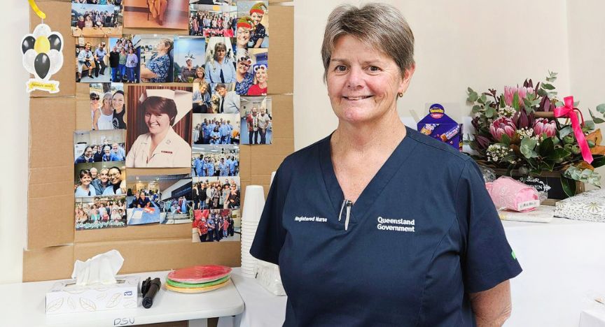 A registered nurse in a Queensland Government uniform stands in front of a collage of photos and a table with flowers and a cake.