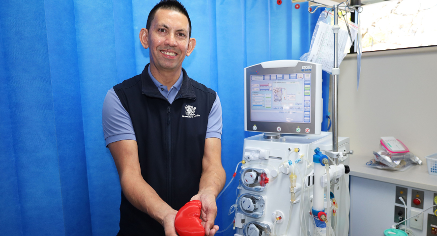 A person in a healthcare uniform holds a red heart model next to a dialysis machine in a medical setting.