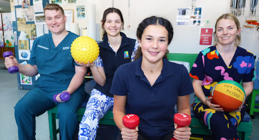 Four physiotherapists holding exercise equipment, including dumbbells and therapy balls, in a clinical setting.