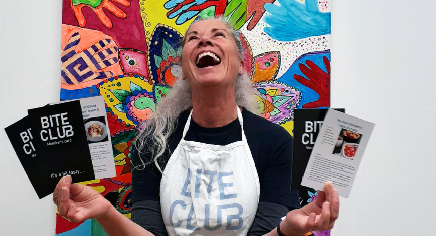 A person wearing a Bite Club apron holds up recipe cards in front of a colorful mural.