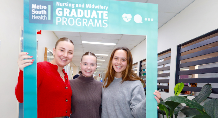 Three people holding a frame that reads Metro South Health Nursing and Midwifery Graduate Programs in a hallway.