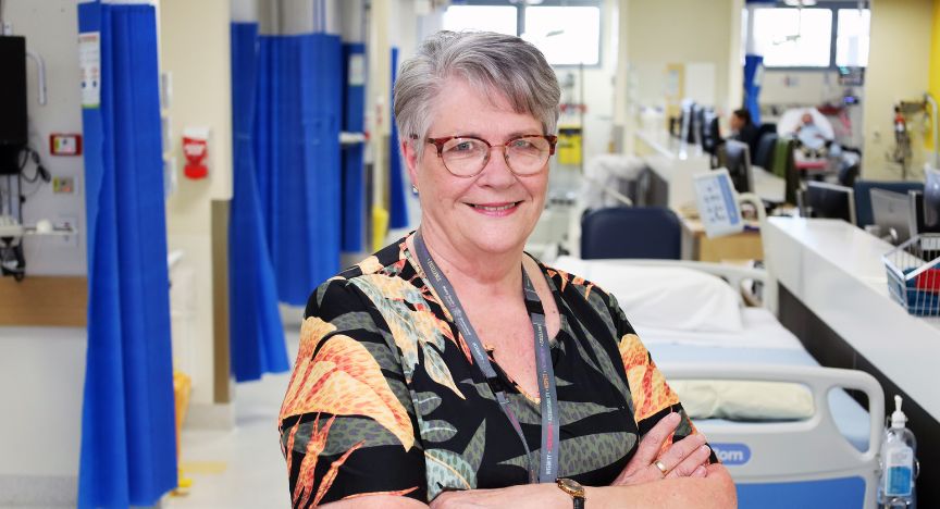 A person stands with arms crossed in a hospital ward, with medical equipment and beds visible in the background.