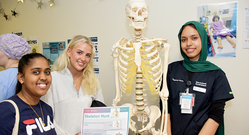 Three people stand next to a skeleton model at a hospital event. A sign titled Skeleton Hunt is visible.