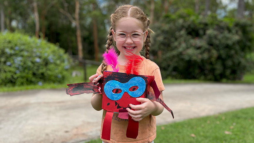 A young girl smiling and holding a decorated bucket called a Mozzie Monster, wearing casual clothes and standing outside with green trees and bushes in the background.