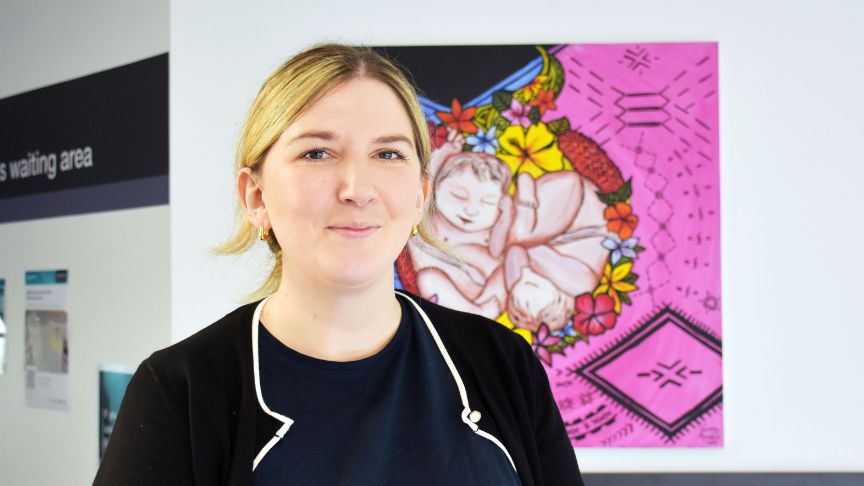 Headshot of female healthcare worker posing in front of a brightly coloured pregnancy canvas. 