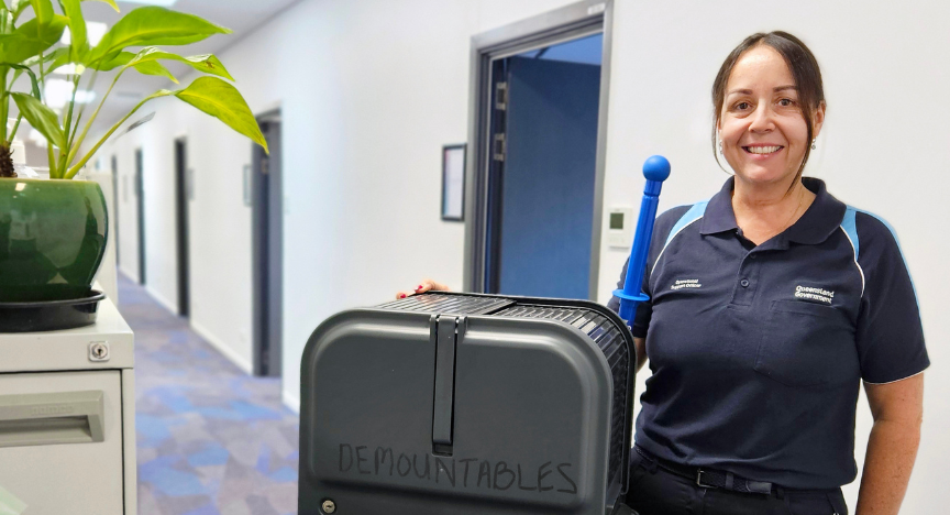 A person in a Queensland Government uniform stands in a hallway, holding a cleaning cart labeled DEMOUNTABLES. A potted plant is on a nearby cabinet.