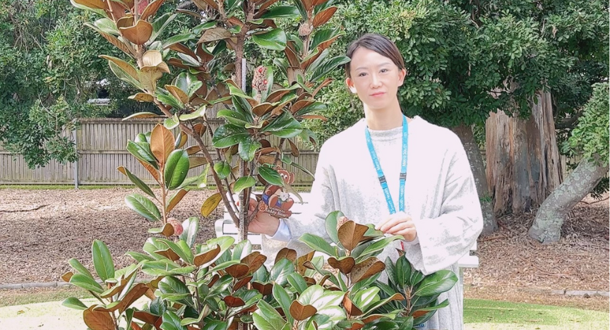 A person holding a First Nations artwork stands beside a small tree in a garden setting.